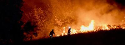 Burning wildfire, orange flames, smoke billowing, dark sky.