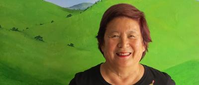 Woman in a field, smiling, holding a camera, trees in background, sunny day.