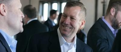 Woman in business attire, holding a microphone, speaking at a podium with a logo.