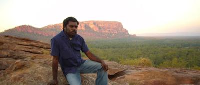 Man in hat, holding a spear, standing in front of a rock formation, landscape.