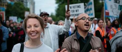 Woman in a white coat, holding a clipboard, smiling, looking at a screen.