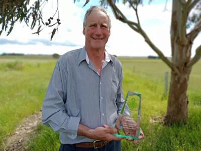Man in a suit, smiling, standing outdoors, trees in background, sunny day.