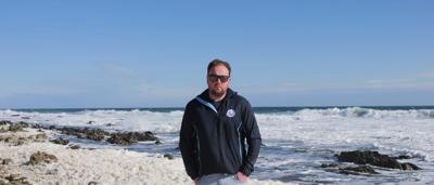 Woman in a white coat, smiling, holding a plant in a greenhouse, surrounded by foliage.