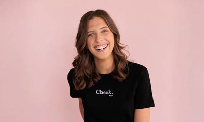 Woman with dark hair smiles, wearing a white shirt, in front of a blurred background.