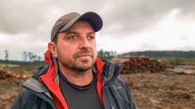 Man in a suit, smiling, standing outdoors, trees in background, sunny day.