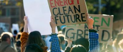 Woman in a white coat, holding a clipboard, smiling, looking at a laptop screen.