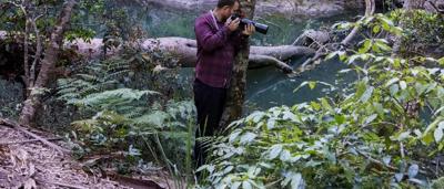 Man in hat, Woody, standing near a river, Broken River, with trees and mountains in background.