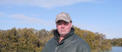 Woman in a white shirt, smiling, holding a camera, outdoors, trees in background.