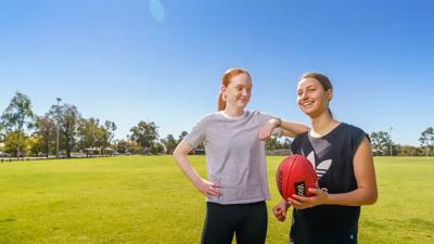 Two women smiling, one with a camera, outdoors, trees in background.