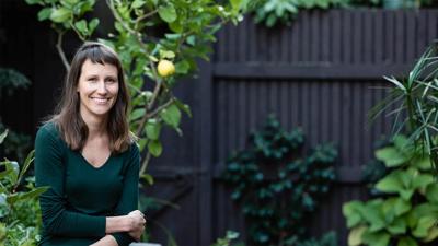 Woman with long hair, smiling, wearing a patterned top, against a blurred background.
