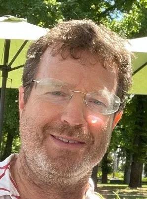Man in hat, holding camera, standing in front of a landscape with trees and a cloudy sky.