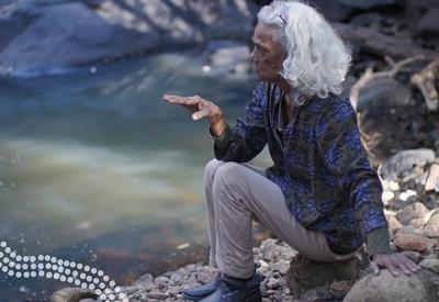 Woman smiling, holding a sign, outdoor setting, trees in background.