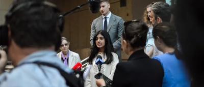 Woman in court, speaking, facing judge, holding documents.